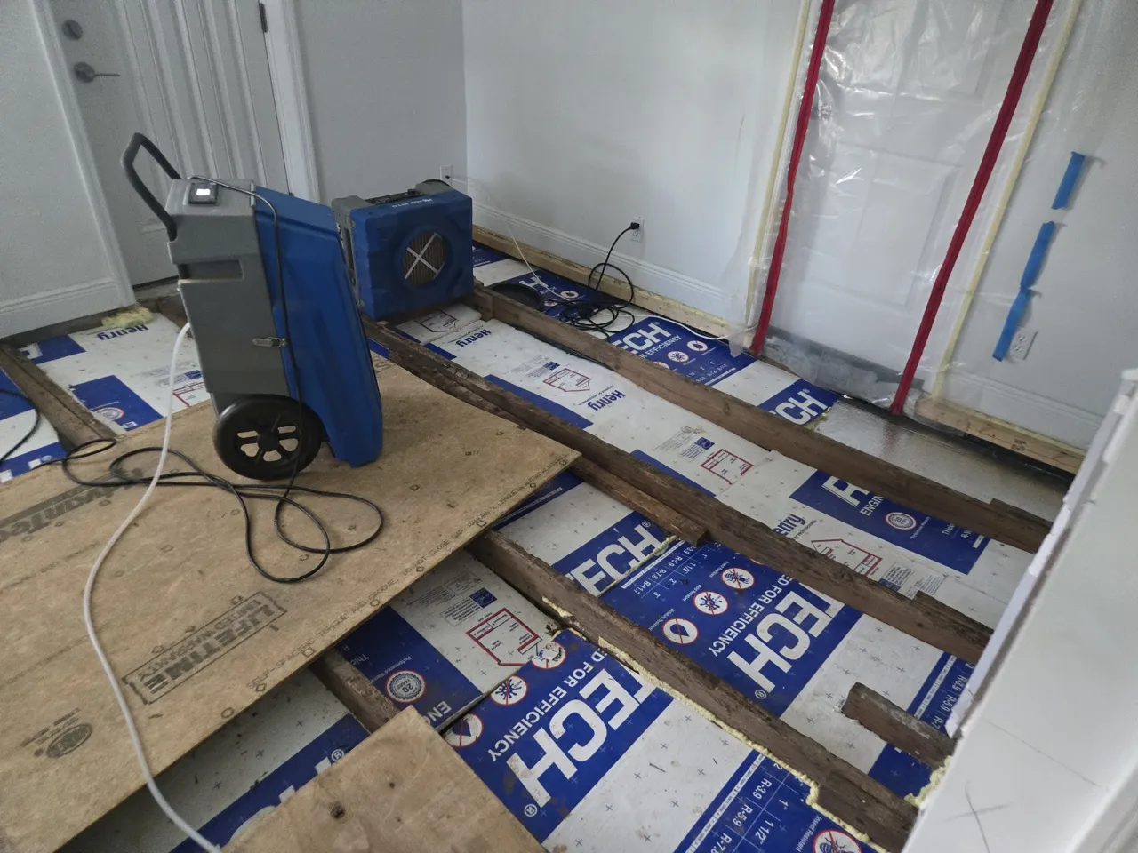Dehumidifier and air mover drying an exposed subfloor inside plastic containment